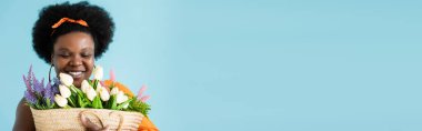 happy african american body positive woman hugging straw bag with flowers isolated on blue, banner