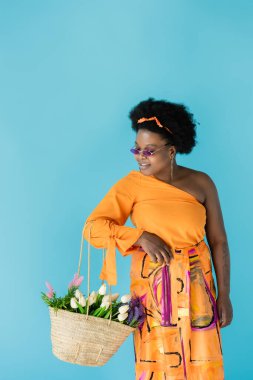 happy african american body positive woman in sunglasses holding straw bag with flowers isolated on blue