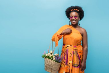 cheerful african american body positive woman in sunglasses holding straw bag with flowers isolated on blue