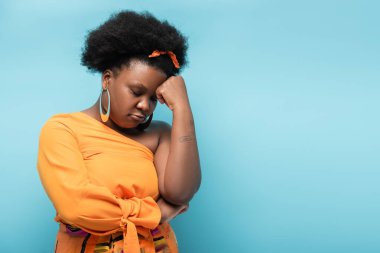 curly african american body positive woman in orange dress and hoop earrings puffing lips isolated on blue