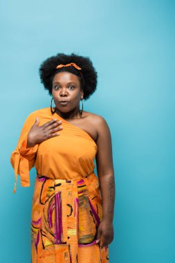 surprised african american body positive woman in orange dress and hoop earrings isolated on blue