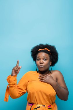 tattooed african american body positive woman in orange dress and hoop earrings pointing with finger isolated on blue