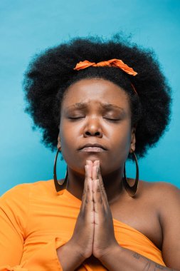 curly african american body positive woman in orange dress and hoop earrings praying isolated on blue