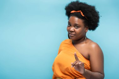 african american body positive woman in orange dress and hoop earrings pointing with finger at camera isolated on blue