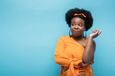 confused african american body positive woman in orange dress and hoop earrings isolated on blue