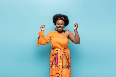 joyful african american body positive woman in hoop earrings and orange dress isolated on blue