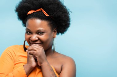 pleased african american body positive woman with tattoo looking at camera isolated on blue