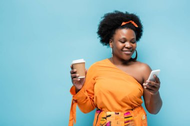 pleased african american body positive woman in hoop earrings holding paper cup and using smartphone isolated on blue