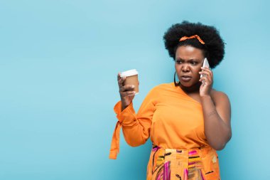 worried african american body positive woman holding paper cup and and talking on smartphone isolated on blue