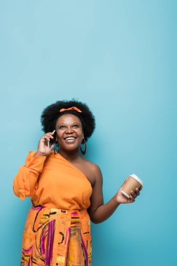 cheerful african american body positive woman in hoop earrings holding paper cup and talking on smartphone on blue