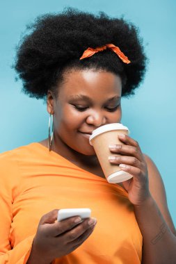 pleased african american body positive woman in hoop earrings holding paper cup and smartphone isolated on blue