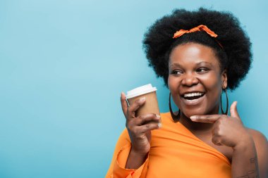happy african american body positive woman in hoop earrings pointing at paper cup isolated on blue