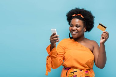 happy african american body positive woman in hoop earrings holding credit card and smartphone isolated on blue