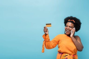 happy african american body positive woman in hoop earrings holding credit card and talking on smartphone isolated on blue