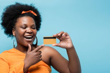 amazed african american body positive woman in hoop earrings pointing at credit card isolated on blue