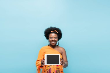 happy african american body positive woman in hoop earrings holding digital tablet with blank screen isolated on blue