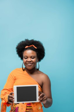 cheerful african american body positive woman in hoop earrings holding digital tablet with blank screen isolated on blue