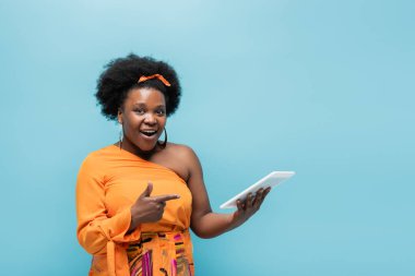 amazed african american body positive woman in hoop earrings pointing at digital tablet isolated on blue