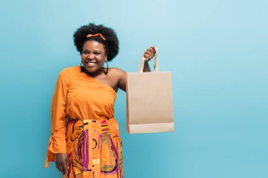 happy african american body positive woman in orange dress holding shopping bag isolated on blue