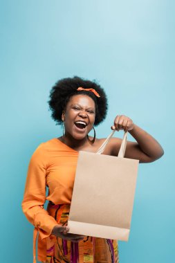 amazed african american body positive woman in orange dress holding shopping bag isolated on blue