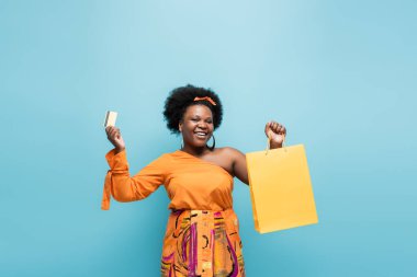 happy african american body positive woman in orange dress holding shopping bag and credit card isolated on blue