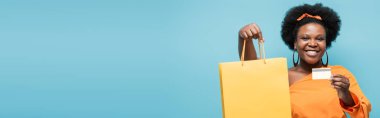 cheerful african american body positive woman holding shopping bag and credit card isolated on blue, banner