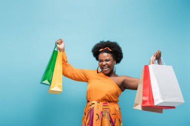 excited african american body positive woman holding shopping bags isolated on blue