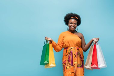 happy african american body positive woman holding shopping bags isolated on blue