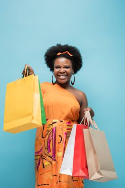 joyful african american body positive woman holding shopping bags isolated on blue