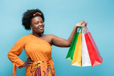 happy african american body positive woman holding shopping bags and posing with hand on hip isolated on blue