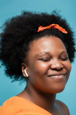 portrait of pleased african american body positive woman in wireless earphone listening music isolated on blue