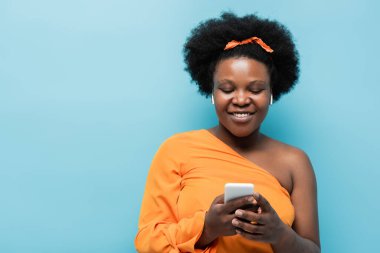 happy african american body positive woman in wireless earphones using smartphone isolated on blue