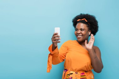 happy african american body positive woman in wireless earphones having video call on smartphone isolated on blue