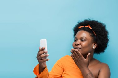 african american body positive woman in wireless earphones having video call and sending air kiss isolated on blue