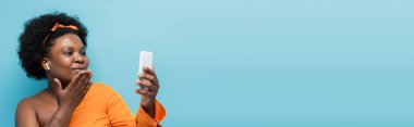african american body positive woman in wireless earphones having video call and sending air kiss isolated on blue, banner