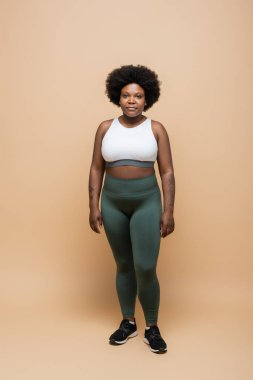 full length of young african american plus size woman in crop top and leggings standing on beige