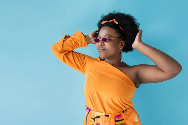 confident african american plus size woman in sunglasses and hoop earrings isolated on blue