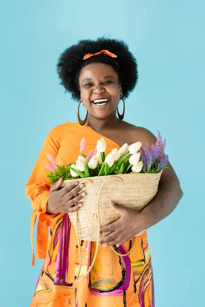 joyful african american body positive woman holding straw bag with flowers isolated on blue