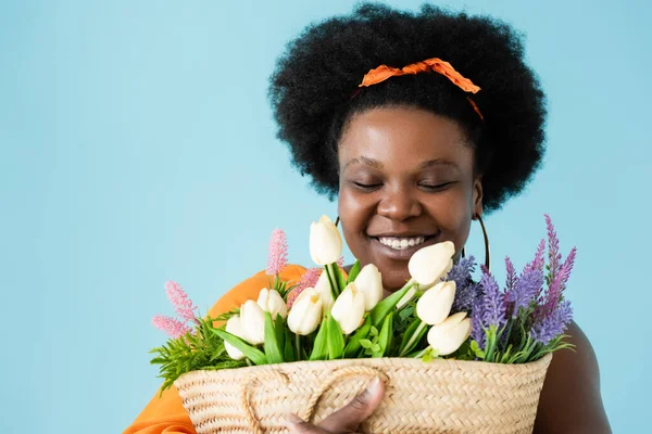 happy african american body positive woman hugging straw bag with flowers isolated on blue