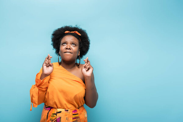 hopeful african american body positive woman in orange dress and hoop earrings with crossed fingers isolated on blue