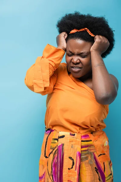 curly african american body positive woman in orange dress and hoop earrings having headache isolated on blue