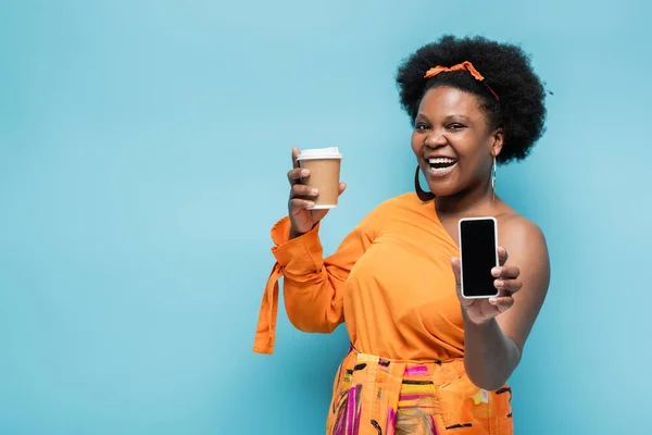 happy african american body positive woman holding paper cup and smartphone with blank screen isolated on blue