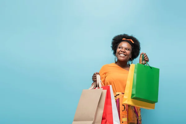 positive african american body positive woman holding shopping bags isolated on blue