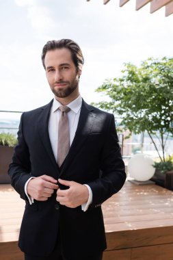 Portrait of groom looking at camera while adjusting jacket outdoors 