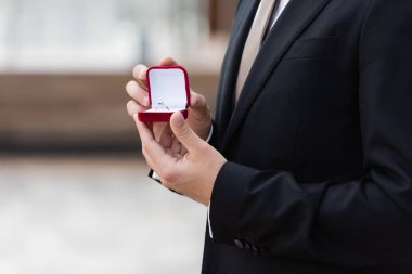 Cropped view of man in elegant suit holding box with engagement ring outdoors 