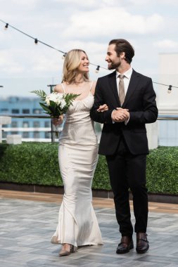 Smiling bride holding bouquet and looking at groom on terrace  