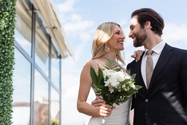 Low angle view of smiling bride with flowers hugging groom on terrace 