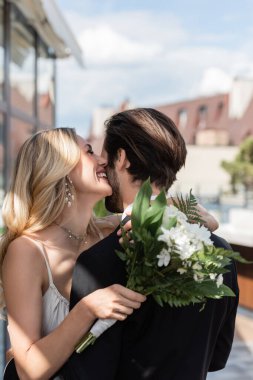 Happy bride holding bouquet and kissing boyfriend on terrace of restaurant 
