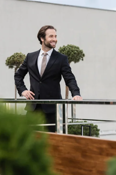 Happy groom in suit standing with hand on hip on terrace of restaurant