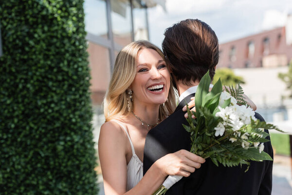 Positive bride with bouquet embracing groom on terrace 